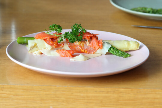 Green and white cooked asparagus wrapped in a piece of ham with cheese, a piece of salmon and parsley on a plate.