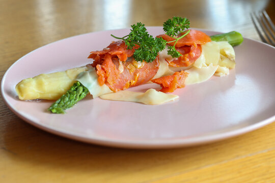 Green and white cooked asparagus wrapped in a piece of ham with cheese, a piece of salmon and parsley on a plate.
