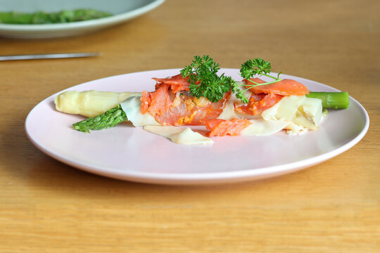 Green and white cooked asparagus wrapped in a piece of ham with cheese, a piece of salmon and parsley on a plate.