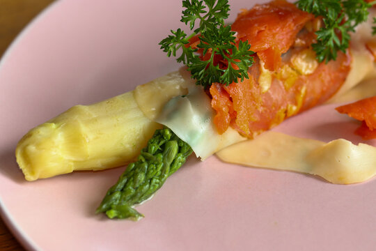 Green and white cooked asparagus wrapped in a piece of ham with cheese, a piece of salmon and parsley on a plate.