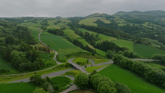 Aerial view of a modern road interchange nestled within the lush, rolling green hills and agricultural patchwork of S o Miguel Island, with a winding rural road and forested areas under a soft