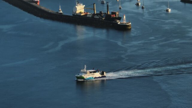 Aerial view of a ferry crossing the waters of Horta harbor on Faial Island, with a cargo ship docked at the pier and various sailboats moored in the background.