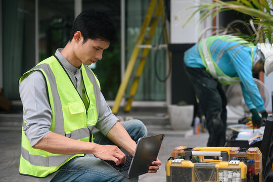 Asian Male Technician in Safety Vest Using Tablet Tablet Outdoors at Construction Site with Colleague Working
