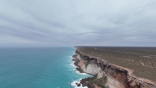 Drone shot flying forward along the Bunda Cliffs, gradually descending toward the hazy ocean under an overcast sky