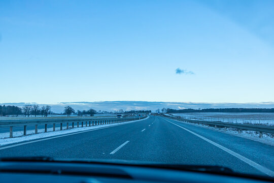 Wide Winter Highway Running Across Frozen Rural Landscape Under Clear Sky. Expansive view of a highway cutting through a frozen countryside with snow-covered edges and a bright blue sky. 