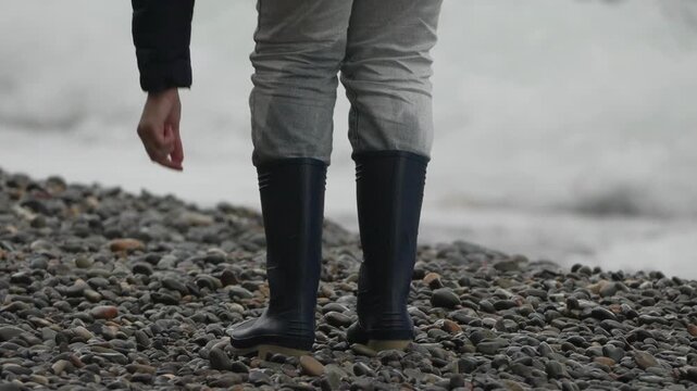 Boots, pebbles, walking person bends down to touch rocks on gloomy shingle beach by ocean surf.
