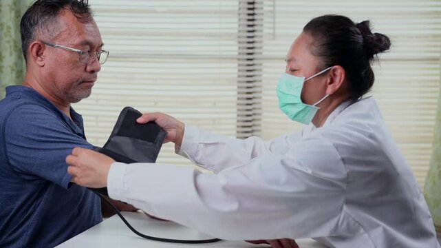 Asian female doctor wearing protective medical face mask measures blood pressure adult male patient sitting inside calm clinic room beside window blinds, gray tablet computer, stethoscope, arm cuff.