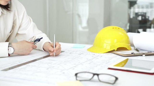 Female architect or engineer working on blueprints using a compass and ruler. Designing building plans. With a yellow hard hat and tablet on the desk