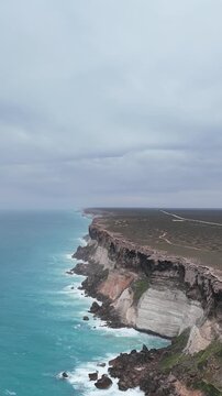 Vertical drone shot flying forward along the Bunda Cliffs, gradually descending toward the hazy ocean with rough white waves under an overcast sky