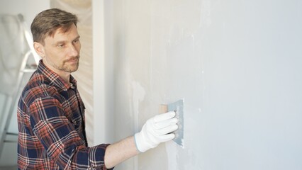 Fototapeta premium Construction worker man wearing gloves and holding a putty knife is applying plaster on a white wall, performing home renovation and improvement