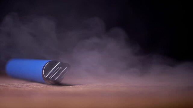 A blue disposable vape pen lies on a wooden table as white vapor curls around the device in a dark setting. Concept of electronic cigarettes, nicotine addiction, and unhealthy habits.