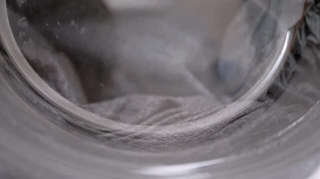 A close-up view of clothes tumbling in soapy water inside the rotating drum of a front-loading washing machine. Concept of household chores, domestic hygiene, and daily appliance use.