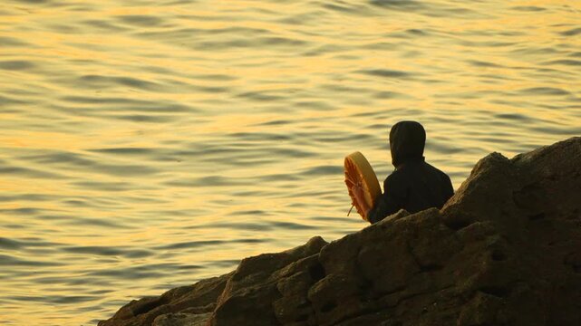 Shaman drumming ocean, hooded figure plays frame drum rhythmically on coastal rocks during mystic golden hour sunset.