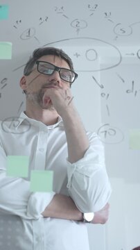 Thoughtful businessman wearing glasses concentrating on complex project plan. Analyzing strategy. Working with sticky notes and flowcharts on a clear glass board