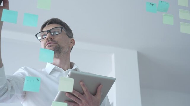 Adult businessman in glasses using a tablet computer while brainstorming and organizing work on a transparent glass board with colorful sticky notes, focusing on strategy and planning