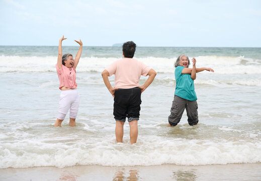 Elderly women exercising on the beach, with gentle waves lapping at their feet. The two women on the sides are stretching