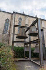 Fototapeta premium Large bronze church bells mounted in a metal frame beside a Gothic stone building with pointed arched stained-glass windows, shrubs, and cloudy sky.