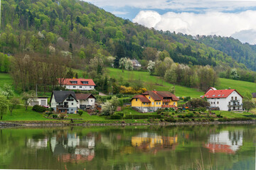 Fototapeta premium Houses with red roofs stand along a green riverside slope, reflected in calm water below forested hills with spring foliage and cloudy mountain scenery.