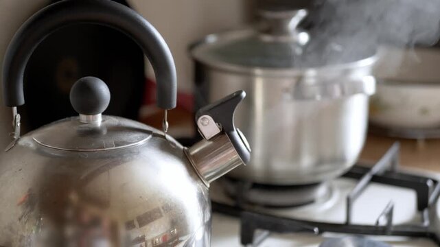 A stainless steel kettle sits on a gas stove burner while a silver pot boils with visible steam rising in a home kitchen. Concept of domestic cooking, morning routine, and boiling water.