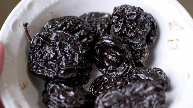 A close-up view of shiny dark prunes packed together in a white ceramic bowl showing their wrinkled texture. Concept of healthy snacking, natural fiber, dried fruit, and organic nutrition.