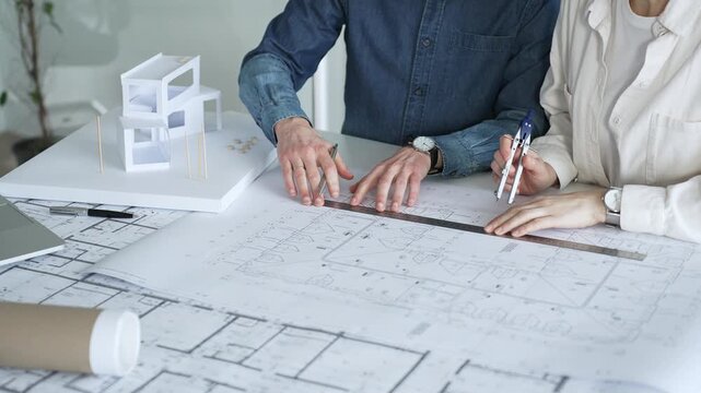 Two architects collaborate over blueprints and a model, using a compass and laptop to measure and plan detailed floor plans, symbolizing teamwork in property development
