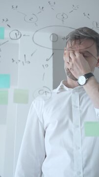 Thoughtful businessman wearing glasses concentrating on complex project plan. Analyzing strategy. Working with sticky notes and flowcharts on a clear glass board