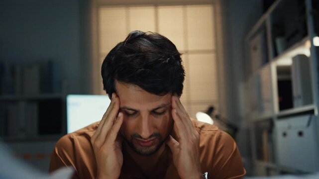 Stressed young Latin businessman having severe headache and migraine at desk in night office. Debt crisis, corporate burnout, late overtime workload, mental health anxiety, business failure concept.
