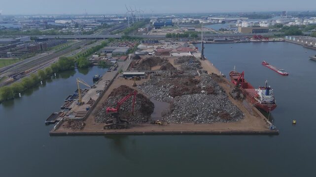 Aerial drone footage circling a large waste and recycling yard located beside the water in an industrial harbor area. Large piles of scrap, debris and waste materials with cranes, ships and port infra