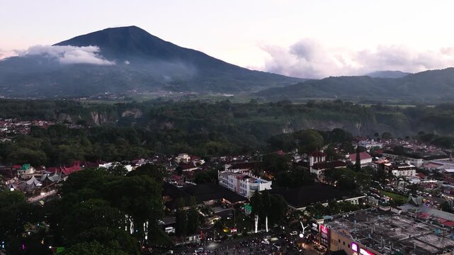 Aerial drone night view of Bukittinggi city lights glowing under a full moon beside Mount Marapi volcano, dramatic nightscape of West Sumatra Indonesia with illuminated urban skyline surrounded