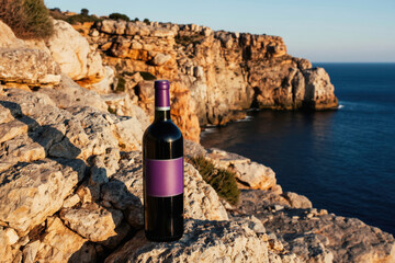 Fototapeta premium Bottle of red wine rests on rocky shore overlooking deep blue ocean. Cliffs rise behind, bathed in warm sunlight, suggesting a perfect vacation drink.