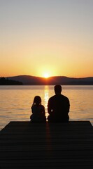 Obraz premium Father and young daughter sit on pier near lake watching sunset. Silhouettes of man and child enjoy peaceful evening, warm orange sky reflection on water.