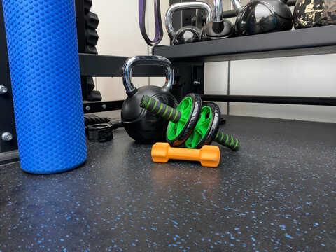 Assorted fitness equipment on a gym floor, including a blue foam roller, green ab wheel, and kettlebells, on a black rubber gym floor. Represents fitness, exercise, and strength training.