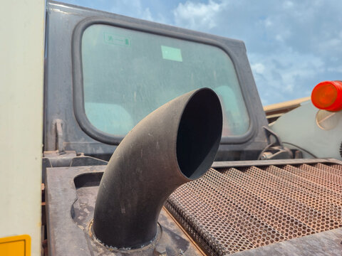 Heavy machinery exhaust pipe close up with cabin window and sky background, industrial vehicle engine ventilation system, construction equipment detail.
