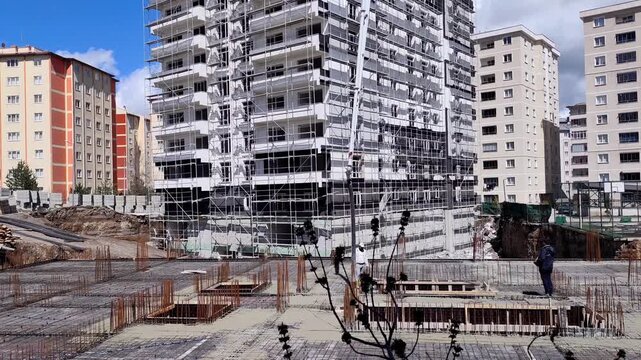 Wide horizontal static shot of a large construction site layout with a deep foundation rebar grid and a high-rise building covered in scaffolding.