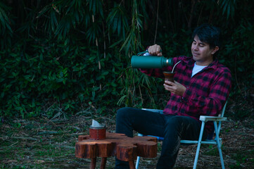 joven con camisa a cuadros roja y negra bebiendo un mate caliente en el parque, expresión de tranquilidad y calma © Daniel