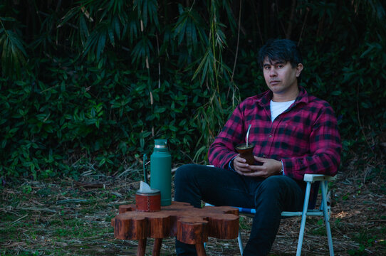 Hombre joven con camisa a cuadros roja y negra mirando a c&aacute;mara y sentado en una silleta en el jard&iacute;n. bebiendo mate con yerba mate.
