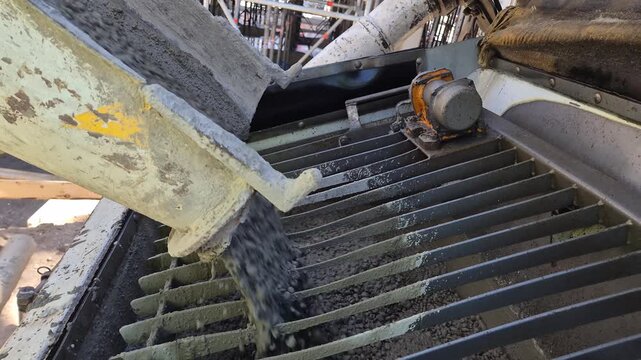 Slow motion side view shot of fresh liquid concrete pouring from a mixer truck chute into the hopper of a concrete pump at a construction site.