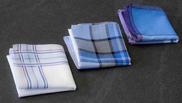 Three different folded men's cotton handkerchiefs in varying blue, white, and plaid patterns  on a textured dark background.