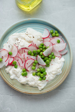 Salad with fresh radish, green peas and torn burrata on a turquoise plate, vertical shot on a light-grey granite background, high angle view
