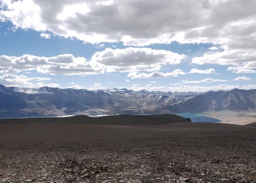 A vast panoramic view of a deep blue alpine lake and barren mountain ranges under a dramatic cloudy sky in Ladakh, Himalayas.