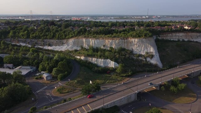 Bluewater cliffs and Dartford Crossing bridge at sunset aerial view. Road with light traffic. Bridge silhouette at distance. Houses build on top of the cliff in golden sunlight.