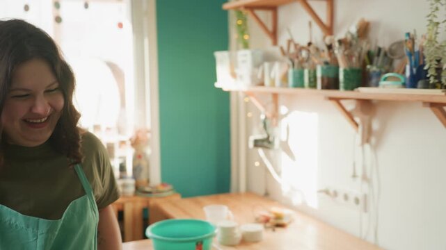 Pottery assistant playing mallet in workshop, green apron, wooden table, jars of glaze on shelves, laughing with rhythmic motion, energetic joyful atmosphere, studio tools visible
