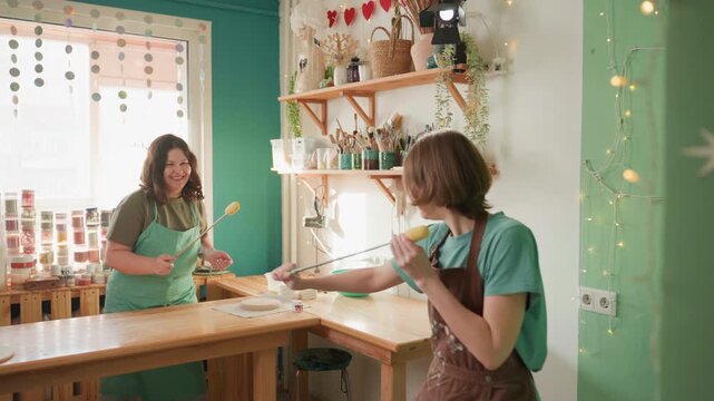 Pottery workshop playful exchange over counter, hands passing brush and sponge. Assistant and owner test colors with jars and bowls on wooden bench, sunlight through window, cheerful demo and casual