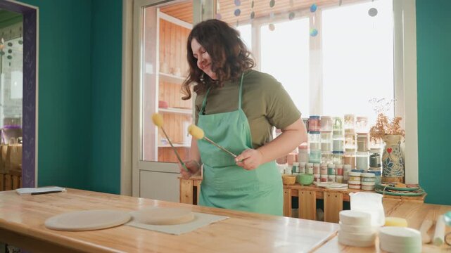 Potter sunlit pottery workshop prepping clay, wearing green apron and smiling while rolling slab on wooden table, arranging jars and tools on nearby shelves, cozy creative atmosphere with glaze