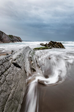 Dramatic long exposure landscape at Itzurun beach in Zumaia showing unique flysch rock formations with silky sea water movement under a cloudy stormy sky in the basque coast