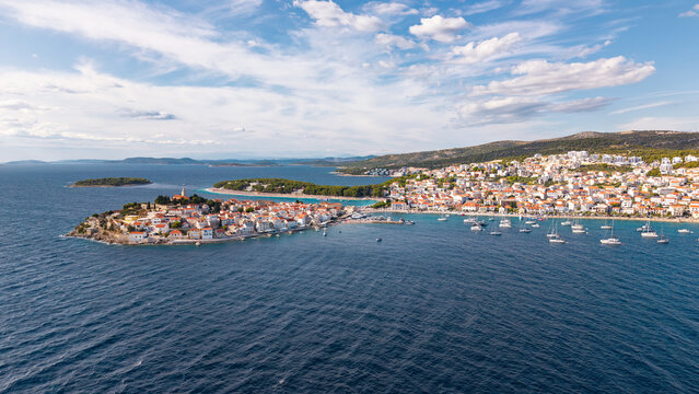 Aerial view of the historic town of Primosten on the Adriatic coast, Croatia, featuring red-roofed houses, a harbor with boats, and the blue sea under a sunny sky