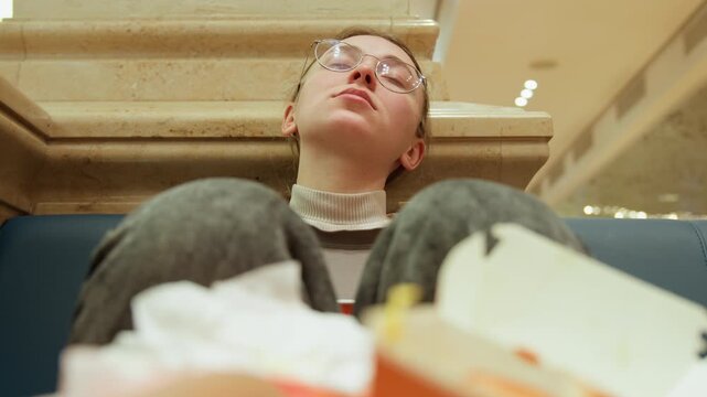 Mall food court exhausted student slumped against bench, glasses reflecting overhead lights, takeout containers and crumpled napkins in foreground, warm ambient lighting, candid portrait showing