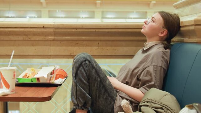 Young white woman resting in booth, tray with burger and fries, backpack at side, gaze lifted to ceiling, pensive expression, soft warm mall lighting, casual sweater and patterned pants, cinematic