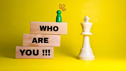 A small green pawn stands on wooden blocks reading "WHO ARE YOU !!!", facing a white chess king against a bright yellow background. © FaizAhmad