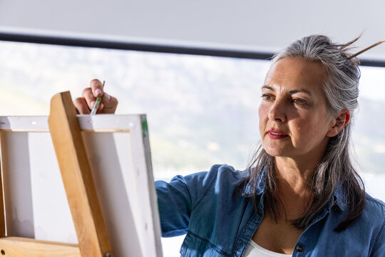 Senior woman painting on canvas at studio with wooden easel holding brush wearing denim, copy space